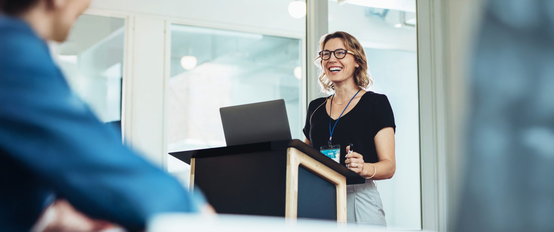 Smiling woman presenting at a professional conference, standing at a podium with a laptop. Highlights inspiration, women in leadership, and confidence in business.