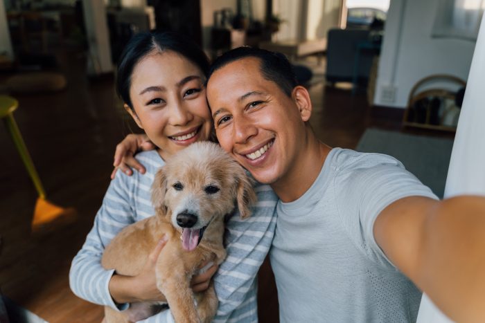 Young adult Asian couple holding a puppy taking a selfie from a phone with home interior in background.
