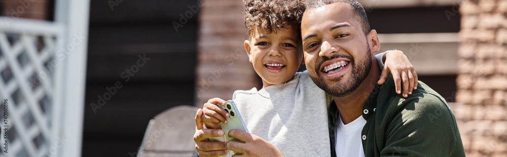 positive african american man in braces and son using smartphone on backyard of house